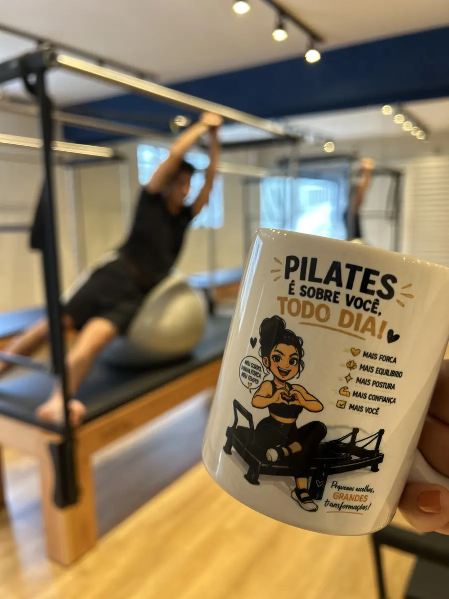 Caneca com a frase "Pilates é sobre você todo dia!" em primeiro plano, com aluno no aparelho ao fundo