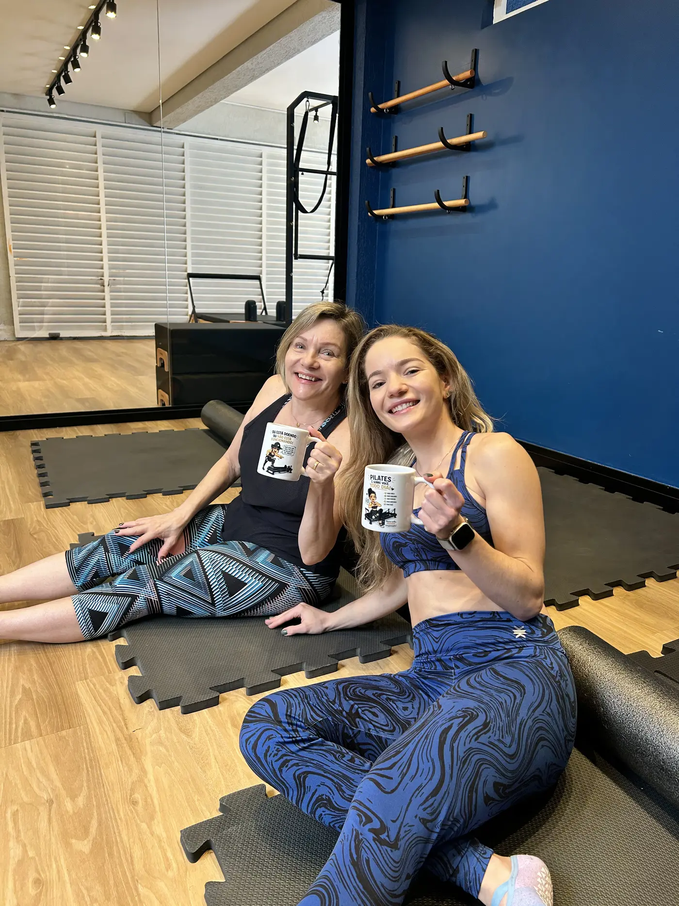 Duas alunas sorrindo sentadas no tatame, segurando canecas do studio